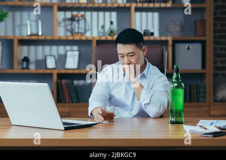 L'uomo d'affari asiatico fallisce, l'uomo disperato beve liquore duro, seduto tardi al tavolo in ufficio, depresso e senza speranza Foto Stock