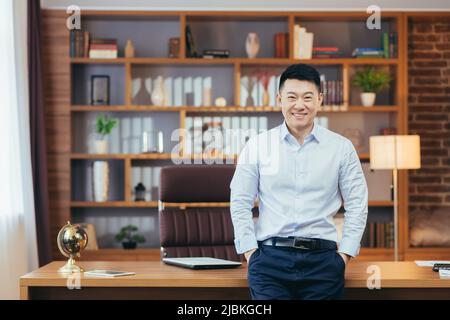 Ritratto di un uomo d'affari di successo, asiatico guardando la macchina fotografica e sorridente, tenendo le mani in tasche pantaloni, uomo in camicia che lavora in un moderno ufficio insur Foto Stock