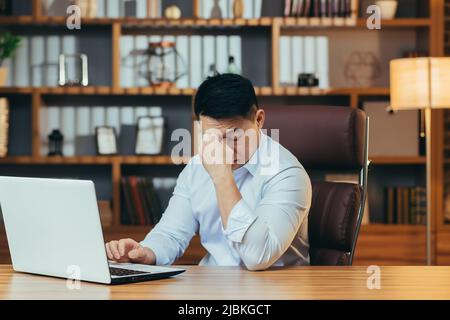 Stanco uomo d'affari asiatico lavora tardi, in un ufficio classico, lavorando su un portatile, pensivo Foto Stock