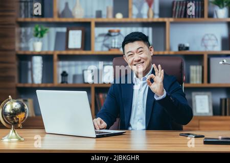 Ritratto di un uomo d'affari asiatico di successo, in un ufficio classico, il capo guarda la macchina fotografica e sorrisi, mostra un segno ok mano Foto Stock