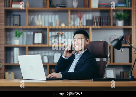 Ritratto di giovane uomo d'affari asiatico bello, avvocato, direttore in abito nero. Parla al telefono, sorride, si siede su una sedia in una scrivania in offi Foto Stock