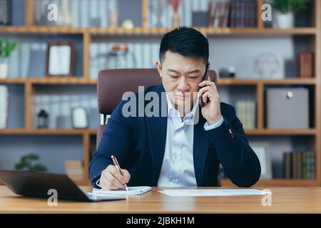 Flusso di lavoro. Giovane bell'uomo asiatico di successo che lavora in ufficio, usando il telefono, parlando al telefono. Direttore, banchiere, avvocato seduto presso la ta Foto Stock