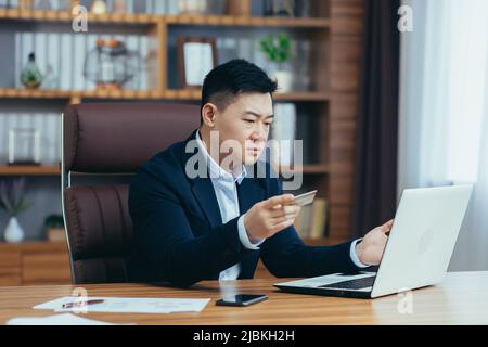 Giovane uomo asiatico di successo banchiere, banchiere che tiene una carta di credito, parlare su un laptop durante una videochiamata, consulenza, vendita. Guardando la macchina fotografica, smile Foto Stock