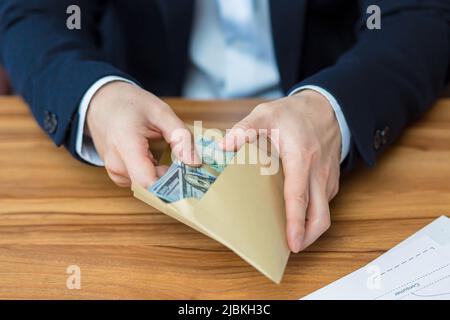 Primo piano le mani della foto del giovane uomo d'affari tiene e apre la busta con soldi, dollari, valuta, tiene in mano al tavolo, mostra, conta Foto Stock