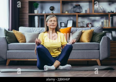 Donna dai capelli grigi più vecchi a casa da sola, stile di vita attivo, yoga, posizione del loto e meditazione Foto Stock