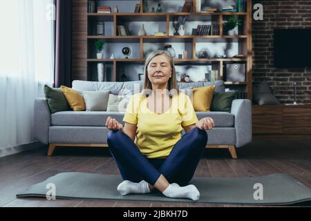 Senior donna dai capelli grigi medita seduta su un tappeto, rilassarsi a casa, fare yoga Foto Stock