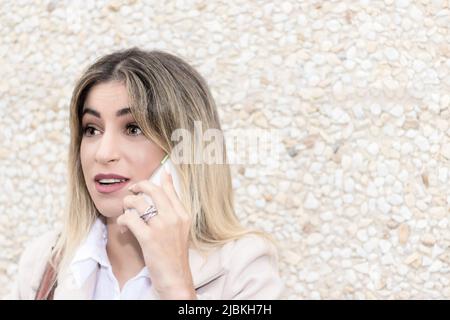 donna bionda che parla sul mobile sulla strada vestita con un cappotto beige e camicia bianca Foto Stock