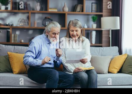 Membro della famiglia di capelli grigi anziani che riceve la lettera o il documento che gioisce seduto a casa. Allegro marito e moglie si ritirees sorridendo mentre si legge il Foto Stock