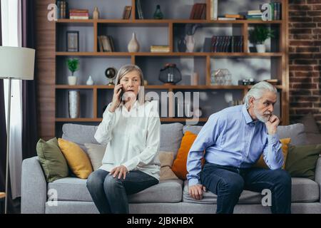 coppia sposata senior, litigante, uomo e donna dai capelli grigi seduti sul divano, moglie infastidita a parlare al telefono Foto Stock