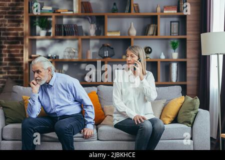 coppia sposata senior, litigante, uomo e donna dai capelli grigi seduti sul divano, moglie infastidita a parlare al telefono Foto Stock