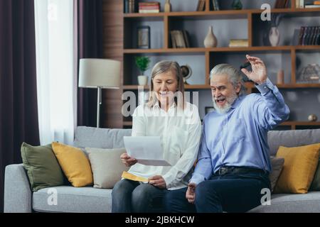 Membro della famiglia di capelli grigi anziani che riceve la lettera o il documento che gioisce seduto a casa. Allegro marito e moglie si ritirees sorridendo mentre si legge il Foto Stock