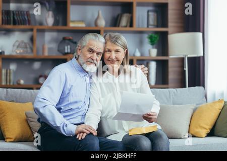 Coppia anziana uomo e donna insieme a casa, ha ricevuto una buona lettera di notizie, felice e sorridente seduto sul divano, guardando la macchina fotografica Foto Stock