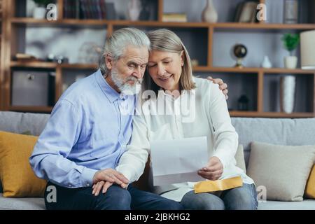 Membro della famiglia di capelli grigi anziani che riceve la lettera o il documento che gioisce seduto a casa. Allegro marito e moglie si ritirees sorridendo mentre si legge il Foto Stock