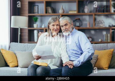 Coppia anziana uomo e donna insieme a casa, ha ricevuto una buona lettera di notizie, felice e sorridente seduto sul divano, guardando la macchina fotografica Foto Stock