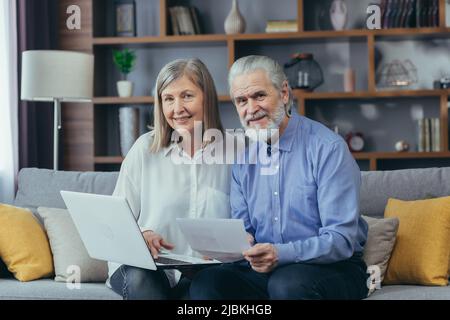 Senior grigio-capelli sposati uomo e donna coppia seduta insieme sul divano a casa, guardando la macchina fotografica e sorridente, carta lavoro con il portatile Foto Stock