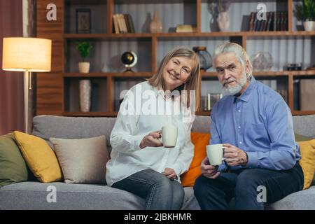 Senior grigio-capelli sposati uomo e donna coppia a casa insieme, rilassarsi insieme in serata, sedersi sul divano e bere bevande calde da Foto Stock