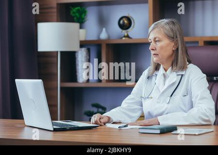 Medico senior con capelli grigi che lavora in un ufficio classico, utilizzando un computer portatile. Responsabile della clinica Foto Stock