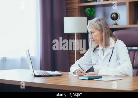 Medico senior con capelli grigi che lavora in un ufficio classico, utilizzando un computer portatile. Responsabile della clinica Foto Stock