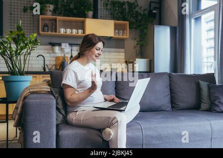 Donna incinta seduta a casa sul divano con un computer portatile, parlando con una videochiamata al medico, si consulta, onde, saldi, sorrisi Foto Stock