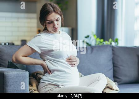Sensazione di malessere. Donna incinta che tiene il suo ventre, sensazione di disagio, dolore. Seduto sul divano, in abiti da casa Foto Stock