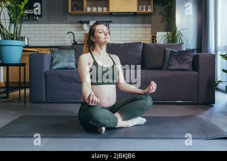 Una donna incinta medita e pratica lo yoga in una posizione di loto a casa Foto Stock