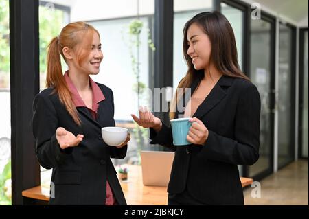 Due belle e di successo donne d'affari asiatiche in tuta formale in piedi in ufficio, avendo un caffè e godere di chiacchierare insieme durante il caffè Foto Stock
