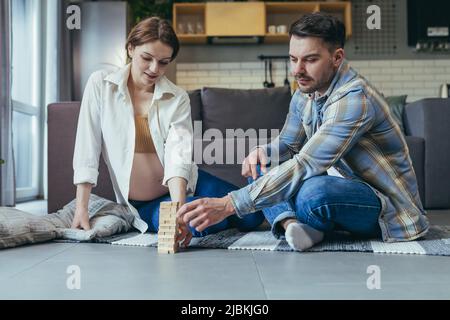 Famiglia giovane. Donna incinta e uomo seduto sul pavimento a casa gioco da tavolo insieme. Fare una torre di barre di legno. Buon divertimento, buon divertimento Foto Stock