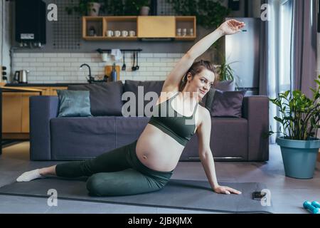 Gravidanza bella donna va in per lo sport a casa, si siede e si trova su un tappeto sportivo in una uniforme sportiva, fa esercizi, è soddisfatto Foto Stock
