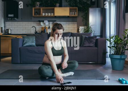 La donna bella incinta va dentro per gli sport nel paese in linea, siede con il telefono su un tappeto sportivo in una uniforme sportiva, fa gli esercizi, è soddisfatta Foto Stock