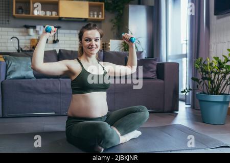 La donna bella incinta entra per gli sport nel paese, siede con i manubri su un tappeto sportivo in una uniforme sportiva, fa le esercitazioni, è soddisfatta Foto Stock