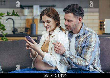 Famiglia giovane seduta sul divano a casa. Donna e uomo incinta. La donna che tiene un telefono nelle sue mani mostra al marito, chiamando insieme genitori, amici. Foto Stock