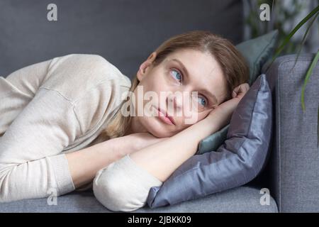 Foto ravvicinata di una donna pensiva di mezza età sdraiata su un divano, guardando fuori dalla finestra a casa Foto Stock