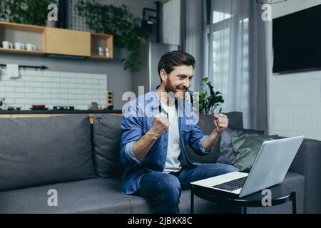 Felice uomo finito il lavoro sul progetto, felice di lavorare al computer, tenendo mano al gesto di vittoria, uomo a casa che lavora con il portatile Foto Stock