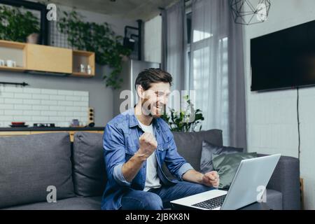Felice uomo finito il lavoro sul progetto, felice di lavorare al computer, tenendo mano al gesto di vittoria, uomo a casa che lavora con il portatile Foto Stock