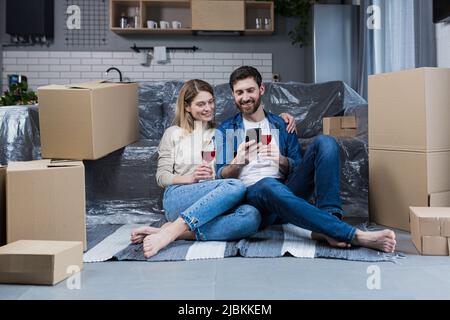 Una coppia sposata innamorata, un uomo e una donna celebrano una festa casalinga, bevono vino da scatole di cartone e scelgono nuovi mobili e app per la casa Foto Stock