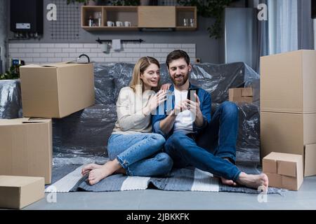 Un uomo e una donna in un nuovo appartamento, una coppia sposata seduta sul pavimento, vicino a scatole di cartone, utilizzando il telefono per lo shopping online, scegliendo furni Foto Stock