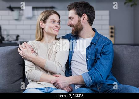 Ritratto di una giovane famiglia felice, coppia. Uomo e donna seduti sul divano, abbracciando, agitando, sorridendo, guardando la macchina fotografica, guardarsi l'un l'altro Foto Stock