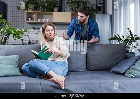 Violenza domestica. Conflitto familiare. Un uomo grida e batte una donna a casa. Donna spaventata chiude le braccia, seduto sul divano. Ha paura Foto Stock