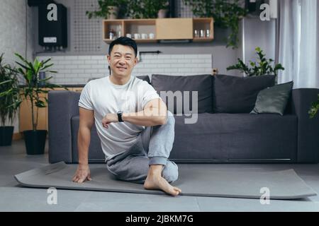 Ritratto di un uomo asiatico felice facendo fitness e fare esercizi mattutini al mattino, uomo seduto su un tappeto sportivo e guardando la macchina fotografica motivare Foto Stock