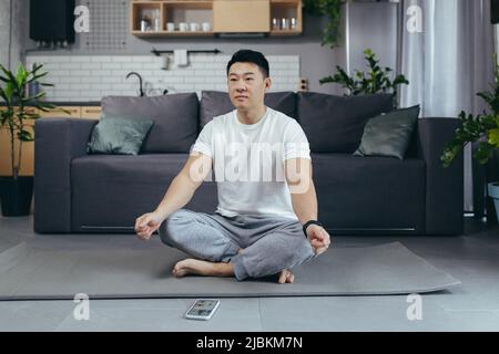 Un uomo a casa è impegnato in meditazione e yoga, un asiatico utilizza un'applicazione mobile al telefono, per il fitness, seduto sul pavimento in una lotus positio Foto Stock