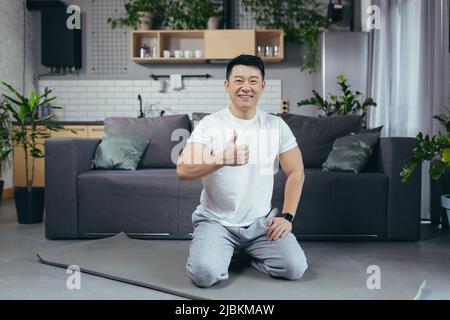 Ritratto di un uomo asiatico felice facendo fitness e fare esercizi mattutini al mattino, uomo seduto su un tappeto sportivo e guardando la macchina fotografica motivare Foto Stock