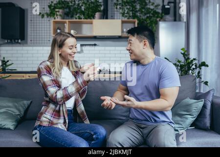 Felice coppia di uomo e donna di razze diverse gioire in gravidanza risultato test, giovane famiglia a casa seduta sul divano e abbraccio Foto Stock