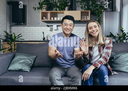 Coppia felice guardando la webcam e sorridendo chattando con gli amici online, uomo e donna di razze diverse, agitando mani gesto di congratulazioni, si Foto Stock