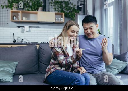 Felice coppia di uomo e donna di razze diverse gioire in gravidanza risultato test, giovane famiglia a casa seduta sul divano e abbraccio Foto Stock