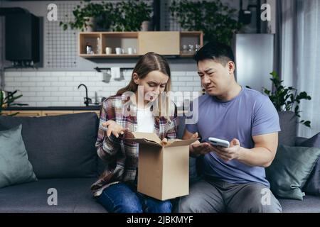 Famiglia multirazziale a casa sul divano deluso di ricevere un pacco, truffato in un negozio online, un uomo e una donna seduta sul divano Foto Stock