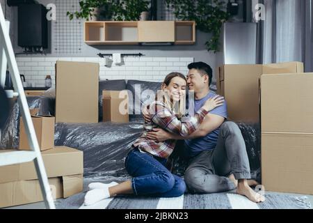 Giovane coppia multirazziale asiatico uomo e donna, in un appartamento appena affittato, abbracciando seduto sul pavimento della casa, scatole di cartone disimballato Foto Stock