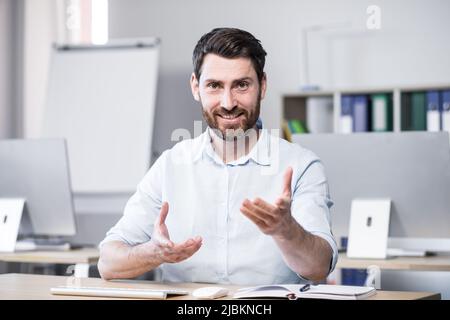 Il giovane bel business coach di successo spiega e racconta con la fotocamera, i gesti con le mani. Seduti su una scrivania del computer in ufficio, guardando i Foto Stock