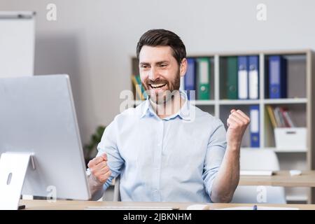 Giovane uomo di successo, lavoratore d'ufficio, manager, freelance si rallegra del risultato del lavoro svolto. Si siede felice al tavolo e al computer, si rallegra, r Foto Stock