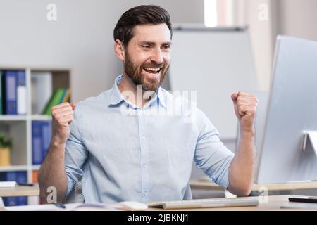 Giovane uomo di successo, lavoratore d'ufficio, manager, freelance si rallegra del risultato del lavoro svolto. Si siede felice al tavolo e al computer, si rallegra, r Foto Stock