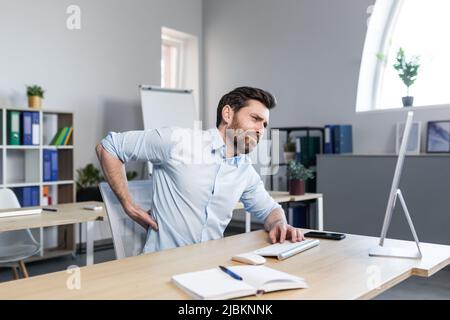 Il giovane uomo stanco, il lavoratore d'ufficio, il freelance che lavora al computer e si siede a una scrivania in ufficio sente dolore alla schiena, massaggia la schiena, si rilassa. Foto Stock